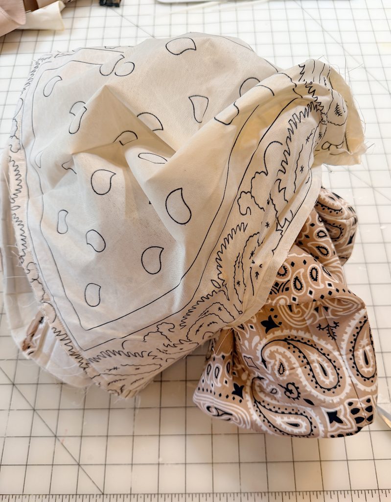 Two patterned bandanas, one white with black outlines and one beige with dark paisley designs, are crumpled on a grid-lined cutting mat.