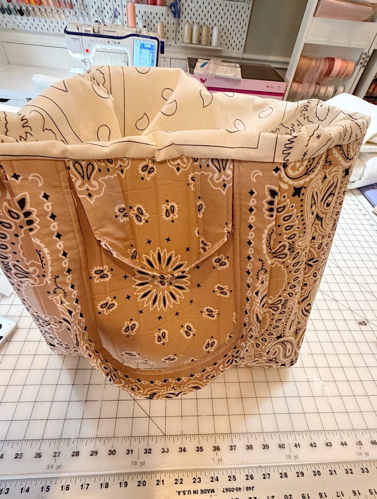 A fabric tote bag with a tan and white bandana pattern is positioned on a grid cutting mat in a craft room with sewing supplies in the background.