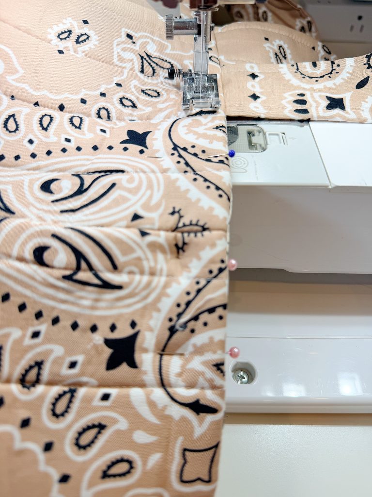 Close-up of a sewing machine stitching beige fabric with a black and white paisley pattern, with fabric secured by pins.