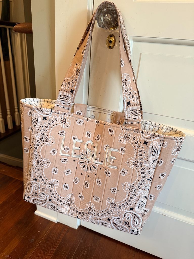 A beige quilted tote bag with a paisley bandana pattern and the name “LESLIE” embroidered on the front, hanging on a glass doorknob.
