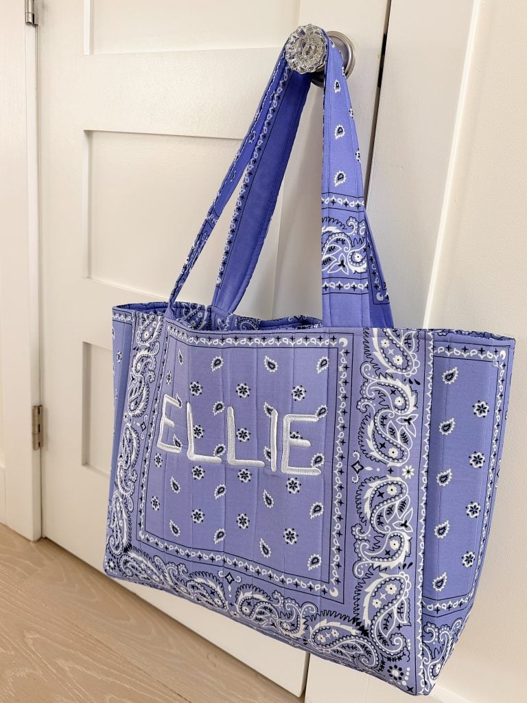 A blue bandana-patterned tote bag with the name "ELLIE" embroidered on the front hangs on a silver door knob.
