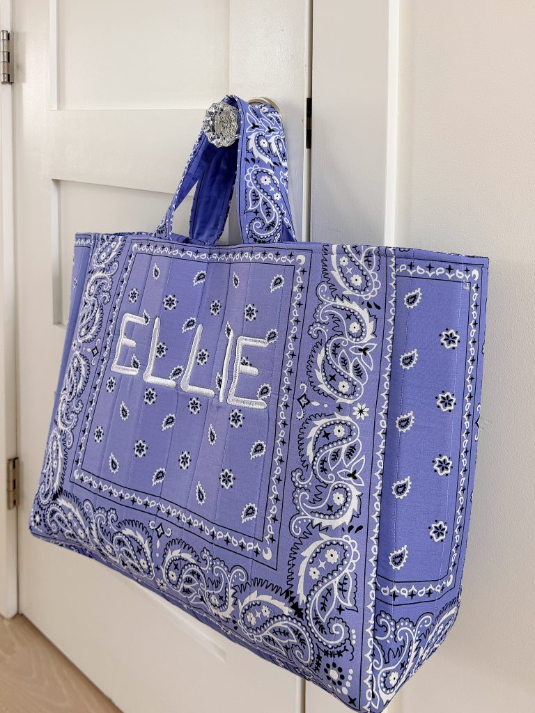A large tote bag made from purple bandana fabric with "ELLIE" embroidered on the front, hanging on a white door handle.