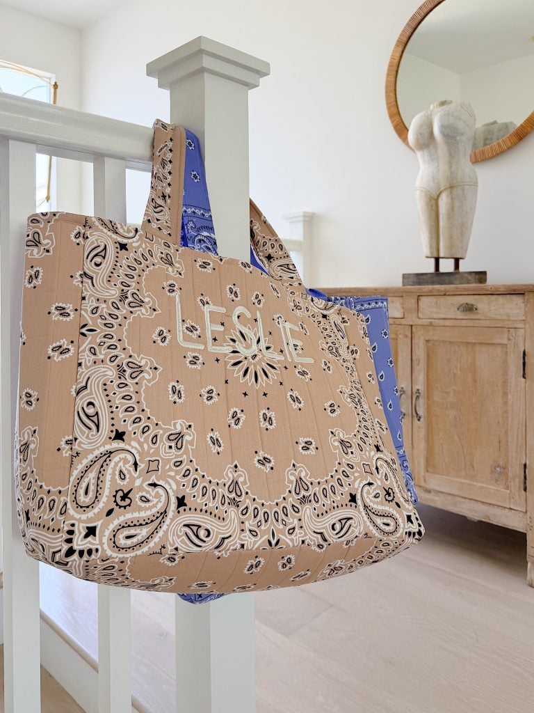 A beige and blue patterned tote bag with the name "LESLIE" hangs on a white stair post in a bright room with wooden furniture and a round mirror.