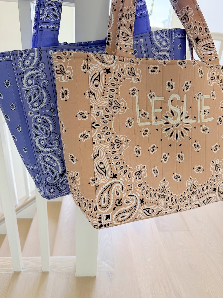 Two patterned tote bags hang on a white railing; one bag is blue, and the other is beige with the name "LESLIE" on the front.