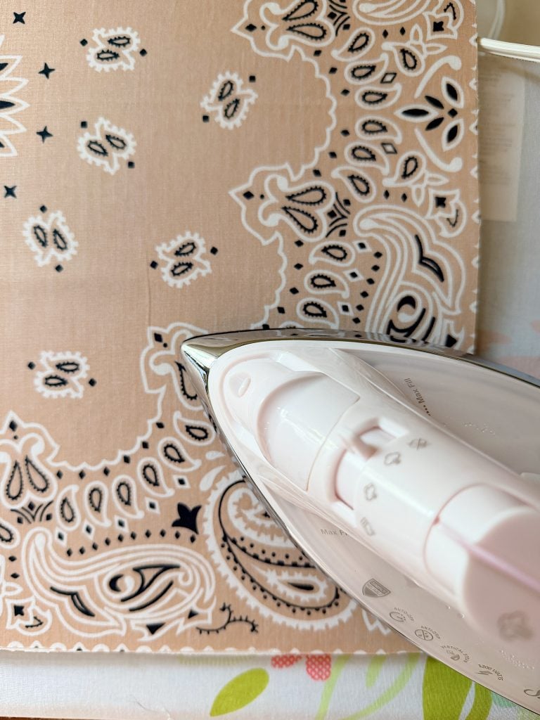 A close-up of a white iron pressing a tan bandana with a black and white paisley pattern.