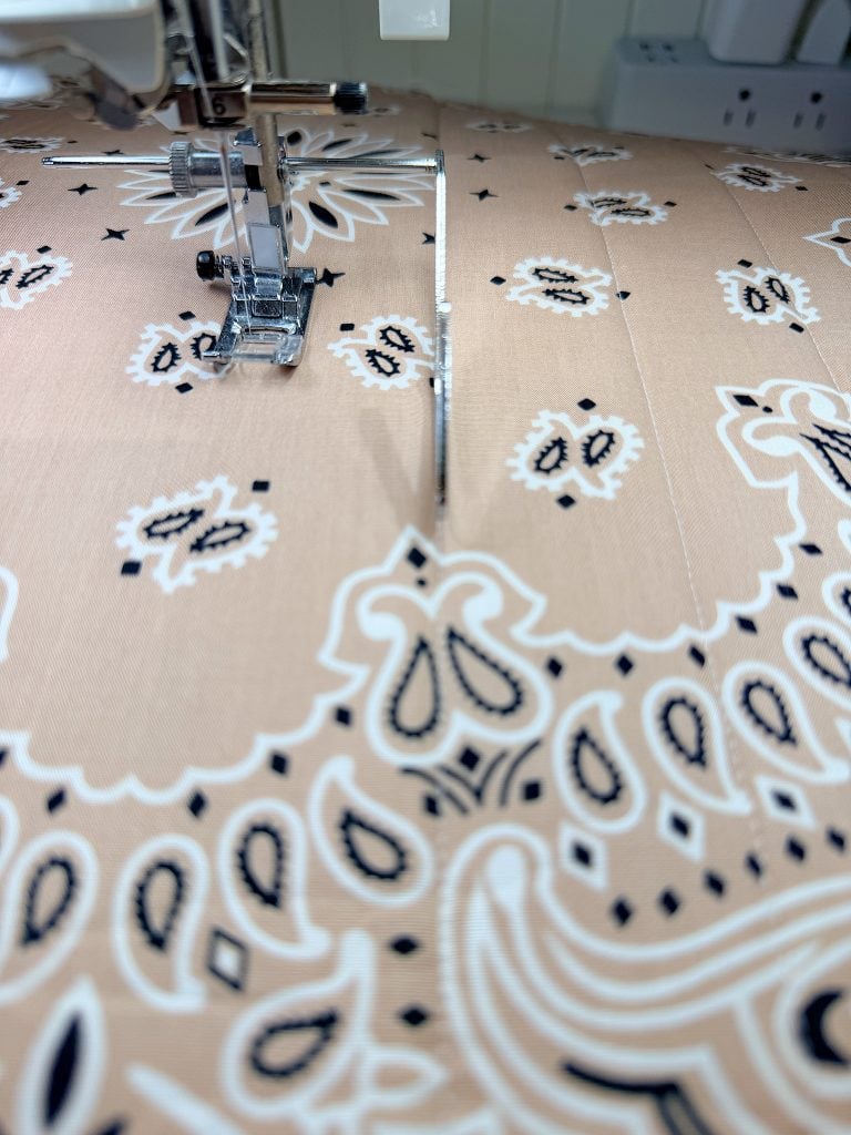 A close-up view of a sewing machine stitching patterned fabric, with the needle in motion and a seam visible in the center.