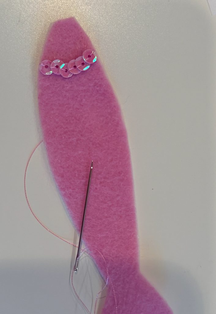 A needle with pink thread rests on a pink felt fish, partially decorated with a row of shiny sequins on its upper side.