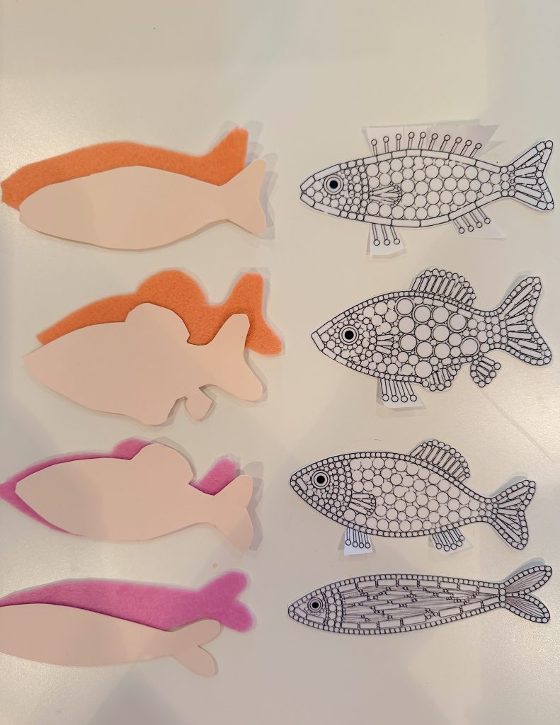 Six paper fish cutouts, three decorated with orange and pink felt, and three with black and white patterned designs, arranged on a white surface.