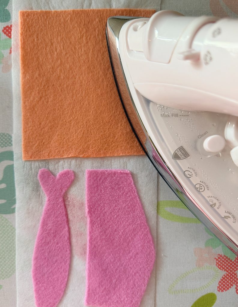 An iron rests on a floral surface next to an orange felt square and two cutouts of pink felt, one fish-shaped and one rectangular.