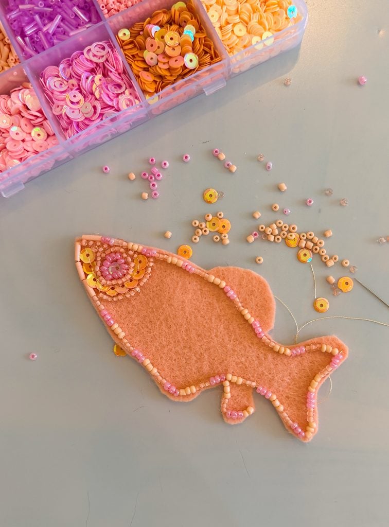Felt fish shape being decorated with pink and yellow beads and sequins, with an open box of sequins at the top left and scattered beads on the table.