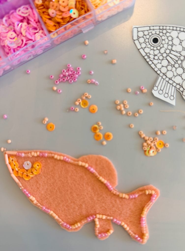 A felt fish is decorated with beads and sequins. More beads are scattered on the table, near a bead organizer and a black-and-white fish outline.