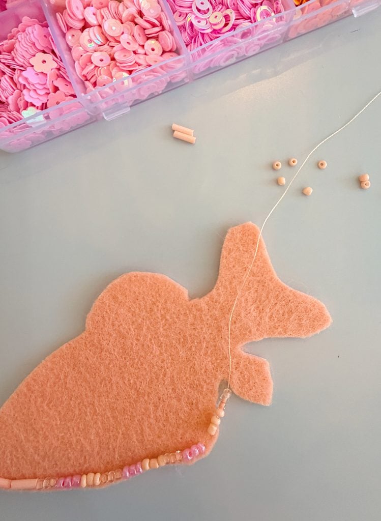 Pink felt cutout being embroidered with small pink beads; a container with pink sequins and beads is visible in the background.