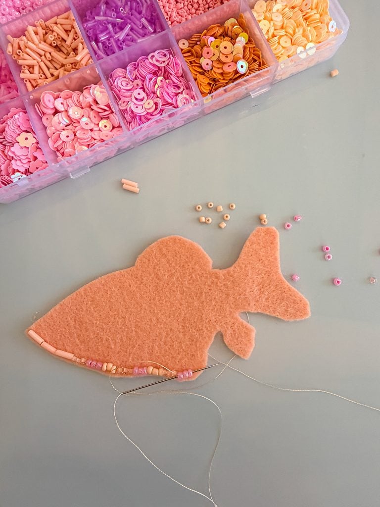 A felt fish shape is being embroidered with beads, with a box of pink, peach, and orange beads above it on a light surface.