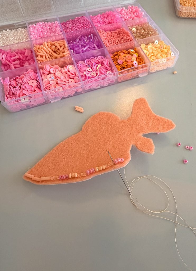 Peach felt carrot shape being decorated with pink and peach beads, needle and thread, and a segmented tray of assorted beads in the background.