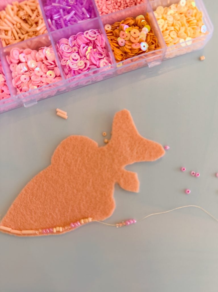 A piece of peach felt shaped like a carrot lies on a table, with a beading kit above and a threaded needle and beads arranged along the carrot’s edge.