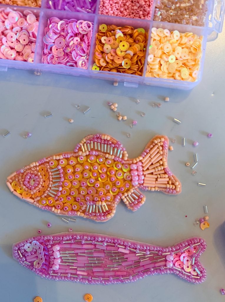 Two fish shapes decorated with beads and sequins in pink, orange, and purple tones are placed near a divided box containing matching craft supplies.