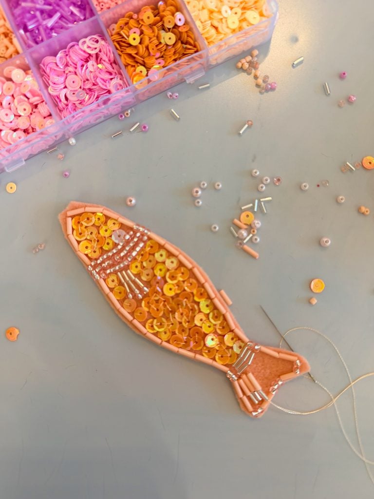 A fish-shaped fabric patch is being decorated with orange and yellow sequins and beads. A box of pink and orange sequins is visible above.