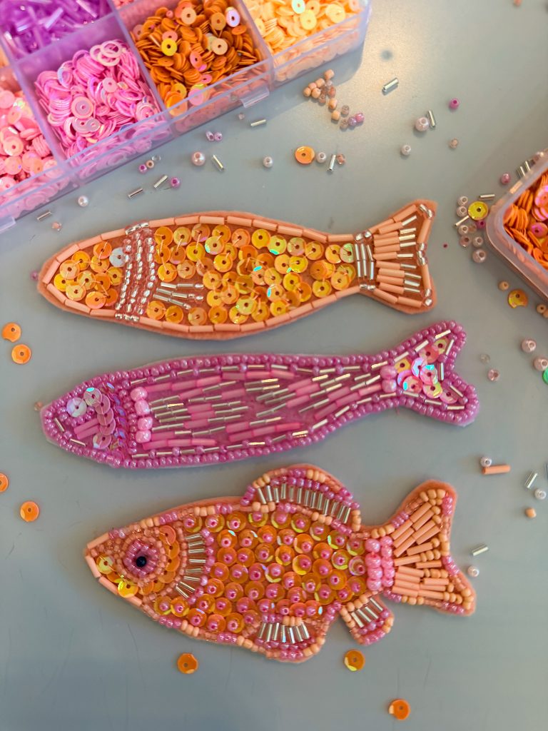 Three fish-shaped crafts decorated with orange and pink beads and sequins are displayed on a table, surrounded by open containers of beads.