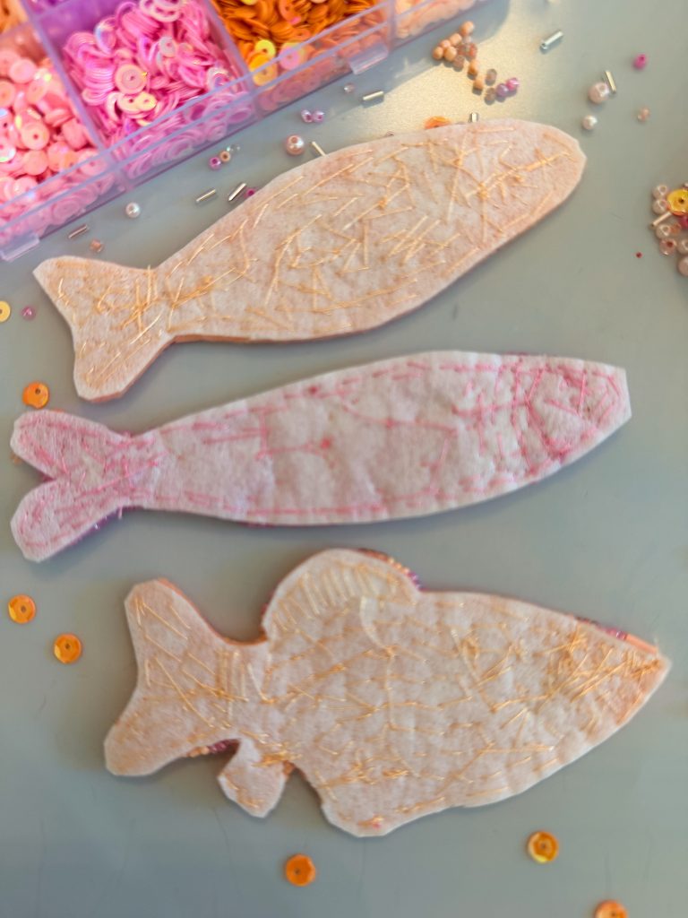 Three felt fish shapes with stitched details are placed on a table, near containers holding various pink and orange sequins and beads.