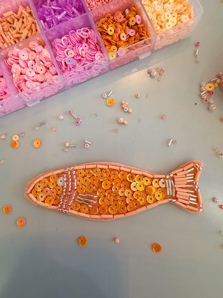 Beads and sequins arranged in the shape of a fish on a table, with a box of organized pink and orange beads in the background.