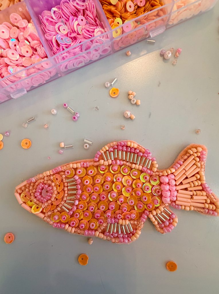 A fish-shaped craft decorated with pink and orange beads and sequins is on a table near an open container of beads.