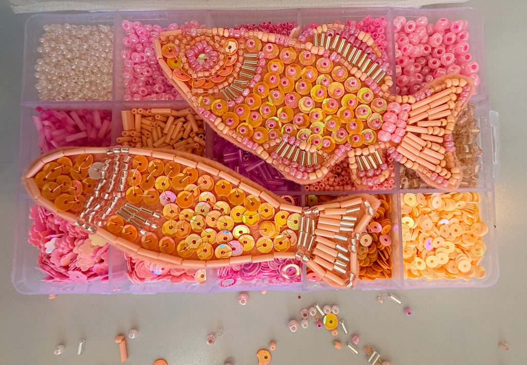 Two fish shapes made of beads, sequins, and small tubes are placed on top of a plastic organizer filled with pink and peach craft beads.