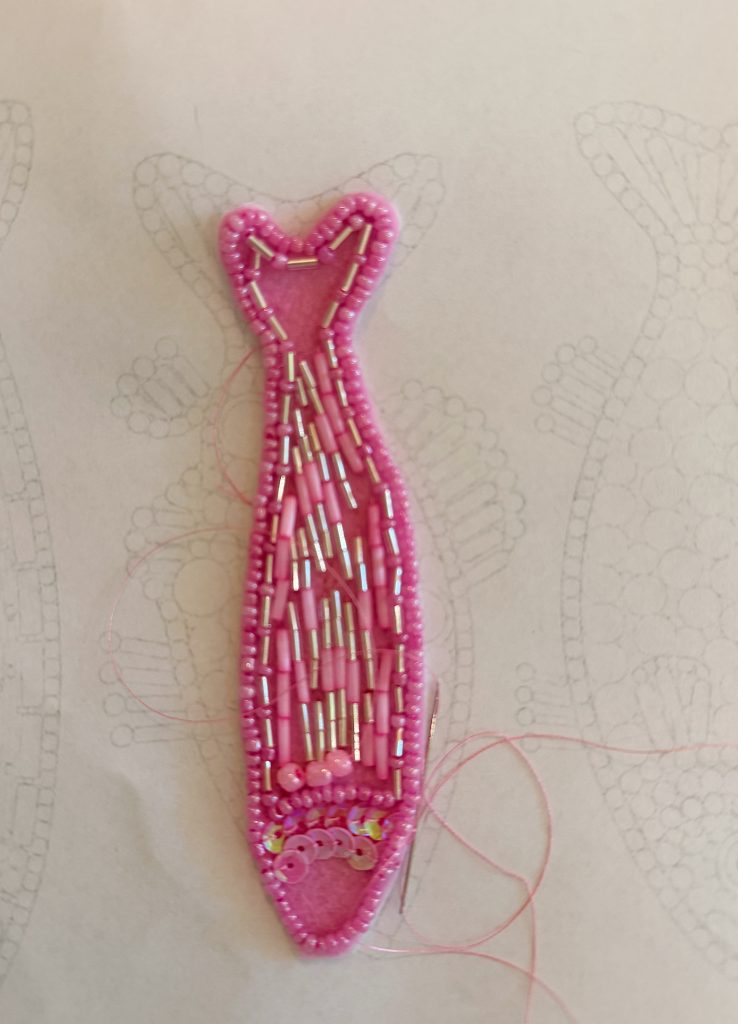 A pink beaded fish shape is partially embroidered on fabric, with pink, silver beads, and sequins. Outlines of more fish are drawn on the background.