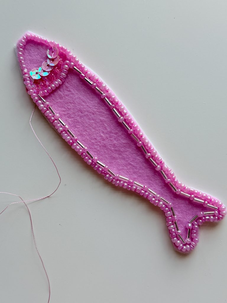 A pink felt fish shape is outlined with beads and sequins, with a needle and pink thread visible on the left side.