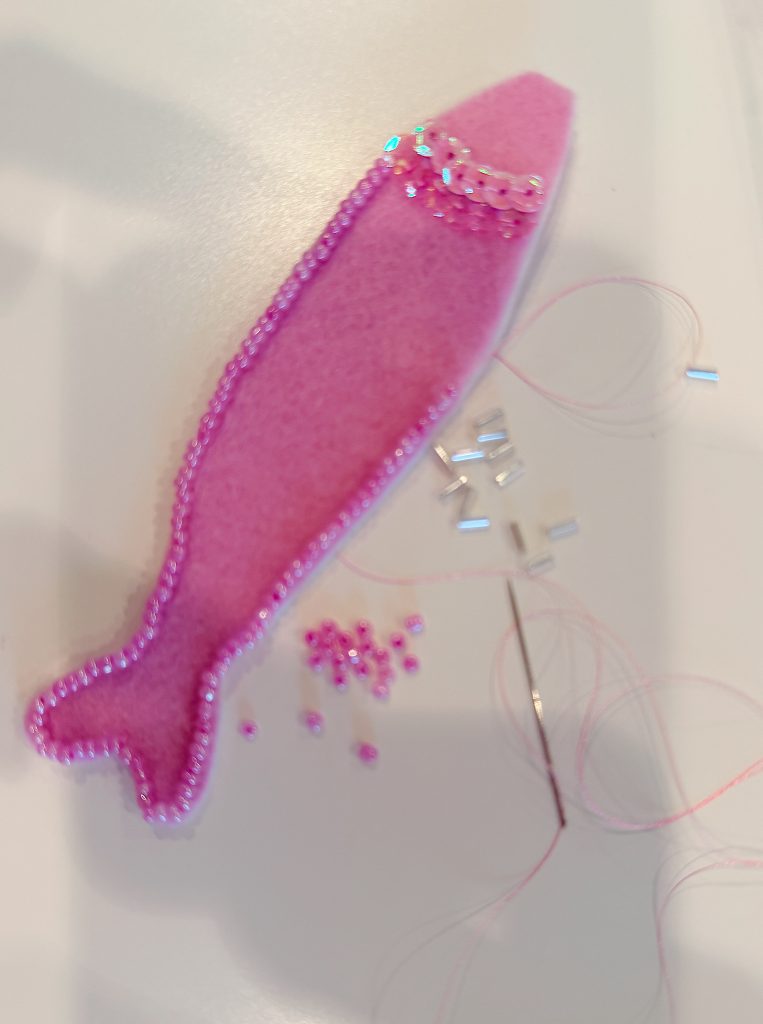 A pink felt fish shape is partially decorated with beads, a needle with thread, and loose beads scattered nearby on a white surface.