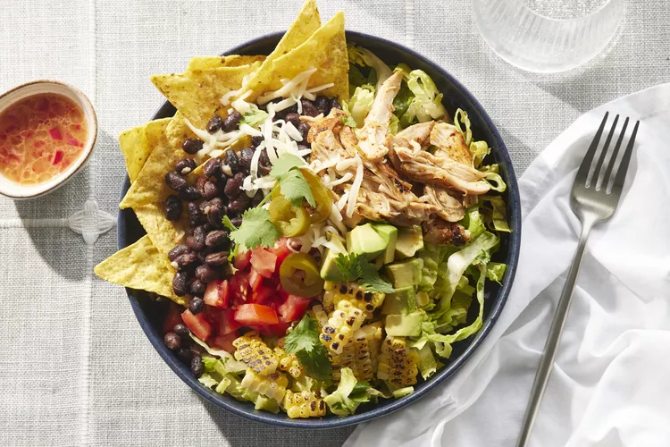 A bowl of salad with shredded chicken, black beans, corn, avocado, tomato, lettuce, tortilla chips, cheese, jalapeños, and cilantro, next to a small cup of dressing and a fork.