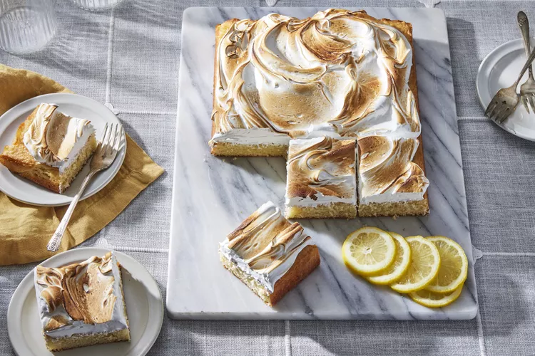 A lemon meringue sheet cake on a marble board, partially sliced, with three lemon slices and two plated servings nearby.
