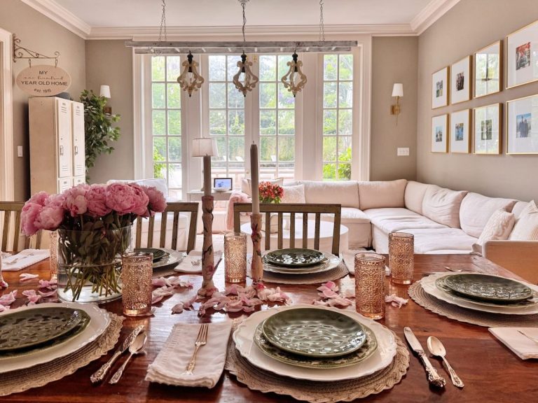 Elegant dining table set with green plates, pink glasses, and floral centerpieces brings summer decor to a bright room with large windows, a beige sectional sofa, and wall art.