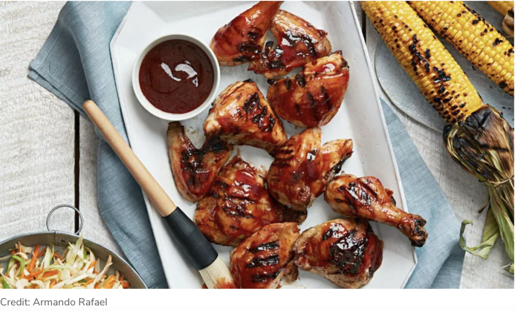 Grilled chicken drumsticks coated with barbecue sauce arranged on a platter with a bowl of sauce, a basting brush, grilled corn, and coleslaw on the side.