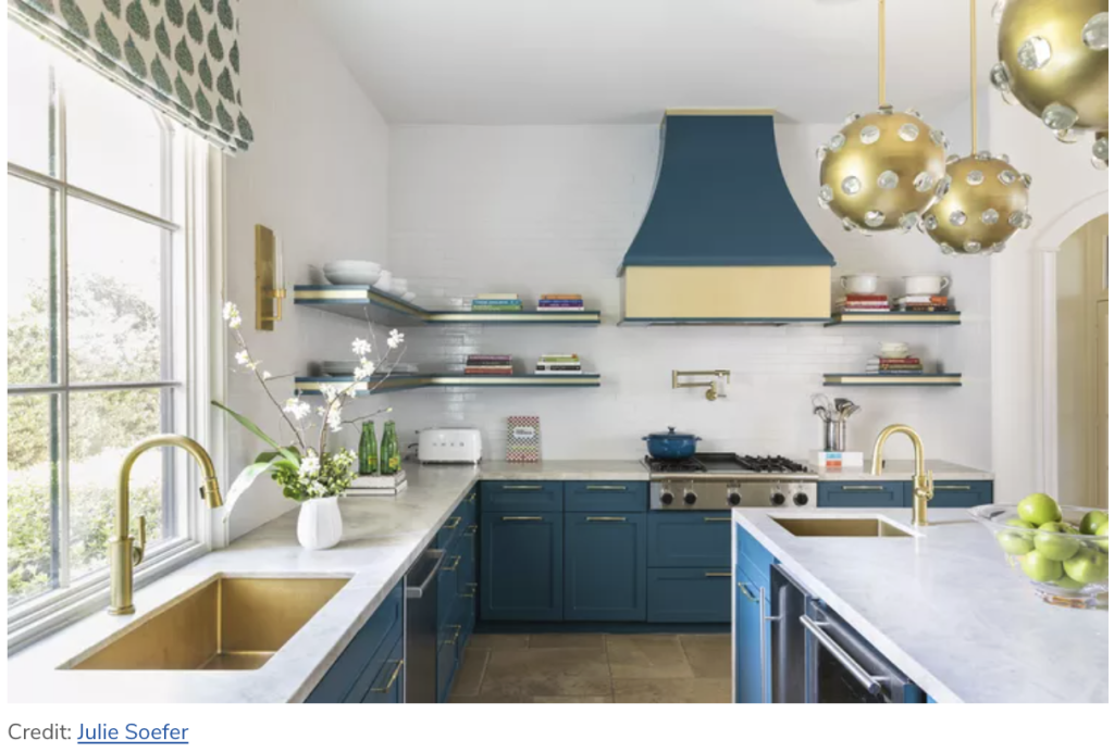 Modern kitchen with blue cabinets, gold fixtures, a blue and gold range hood, marble countertops, open shelves, and globe pendant lights. A window is on the left above a gold sink.