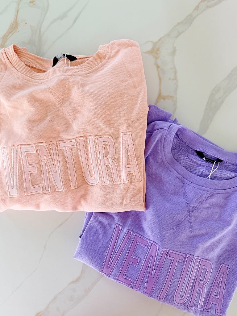 Two crewneck sweatshirts, one peach and one lavender, both with the word "VENTURA" embroidered in large letters across the front, are folded on a marble surface.