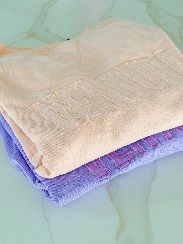 Two folded sweatshirts on a marble surface, one peach and one lavender, both with the word "VENTURA" embossed on the front.