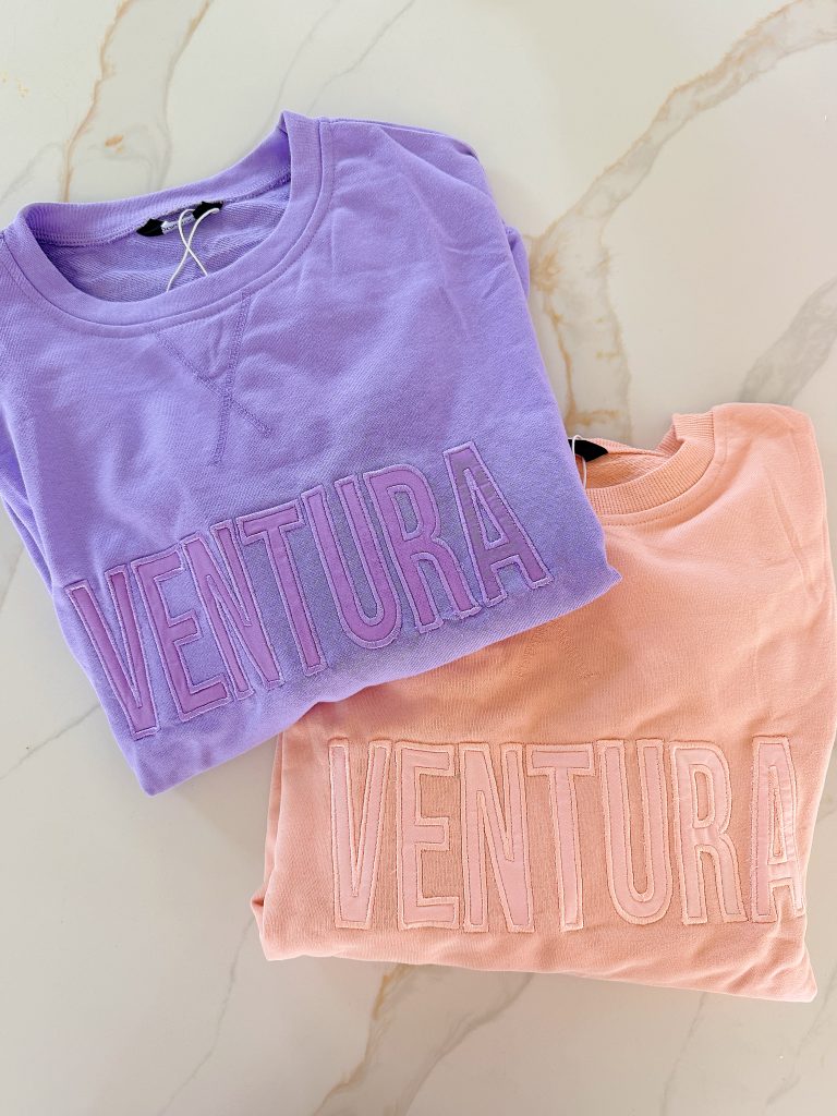Two folded sweatshirts, one lavender and one peach, each with the word "VENTURA" stitched across the front, lying on a light marble surface.