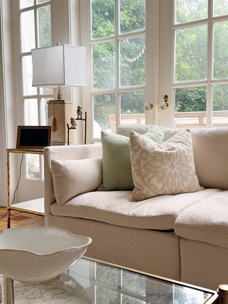 A beige sofa with decorative pillows sits near large glass doors; a side table with a lamp and a digital device is next to it, and greenery is visible outside.