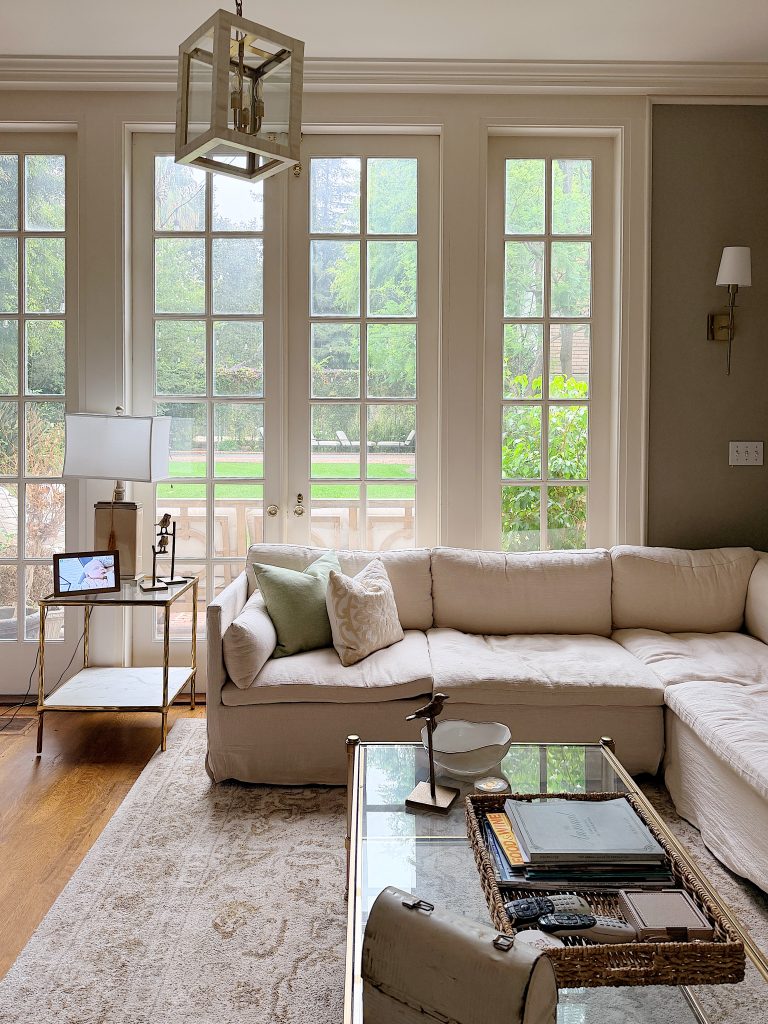 Bright living room with a white sectional sofa, glass coffee table, side table with a lamp and tablet, and large windows overlooking a green backyard.
