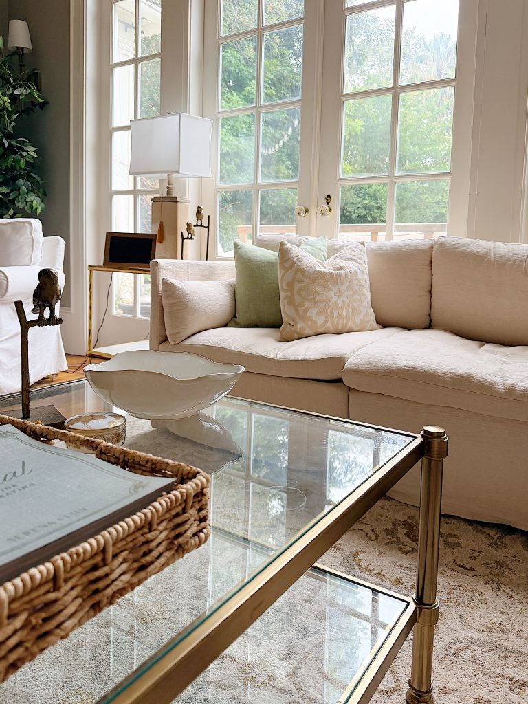 A living room with a white sectional sofa, glass coffee table, wicker tray, decorative bowl, and large windows letting in natural light.