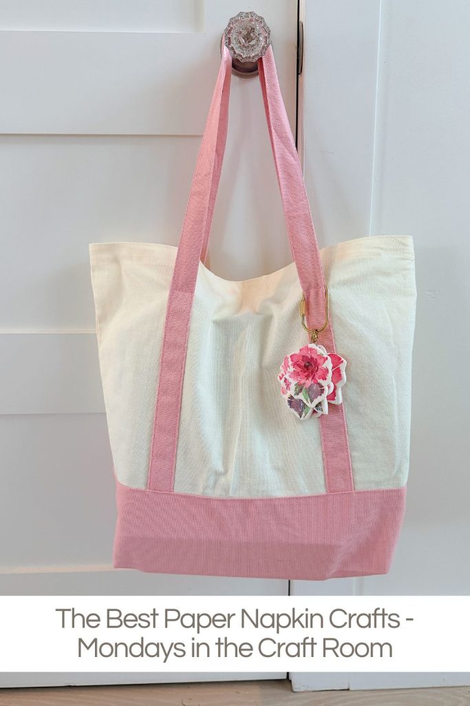 A cream and pink tote bag hangs on a door handle, decorated with a pink paper napkin flower keychain. Text reads: The Best Paper Napkin Crafts - Mondays in the Craft Room.