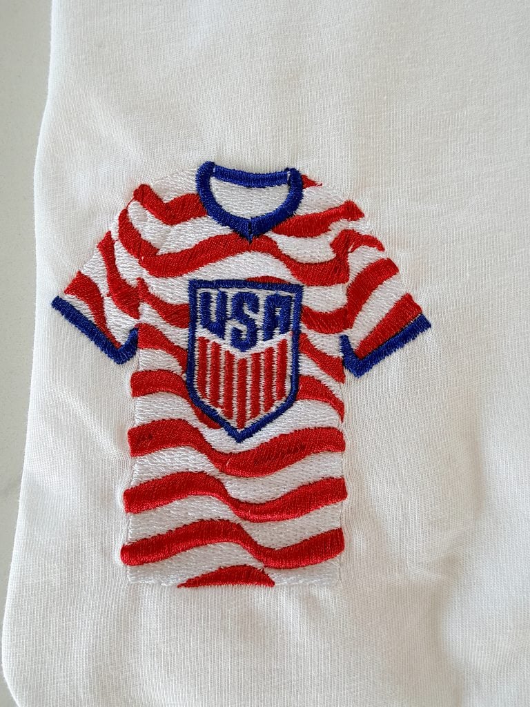 Embroidered red and white striped soccer jersey with blue collar and "USA" shield on a white fabric background.