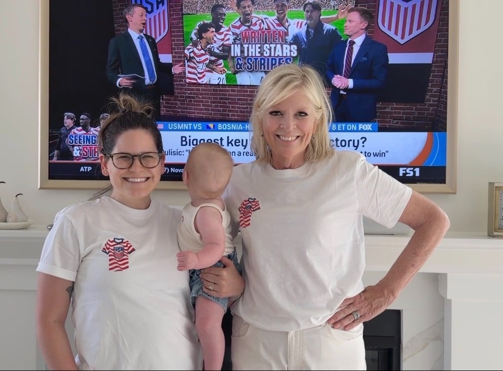 Two women and a baby stand in front of a TV showing a sports broadcast. The women wear matching white shirts with a red, white, and blue heart design.