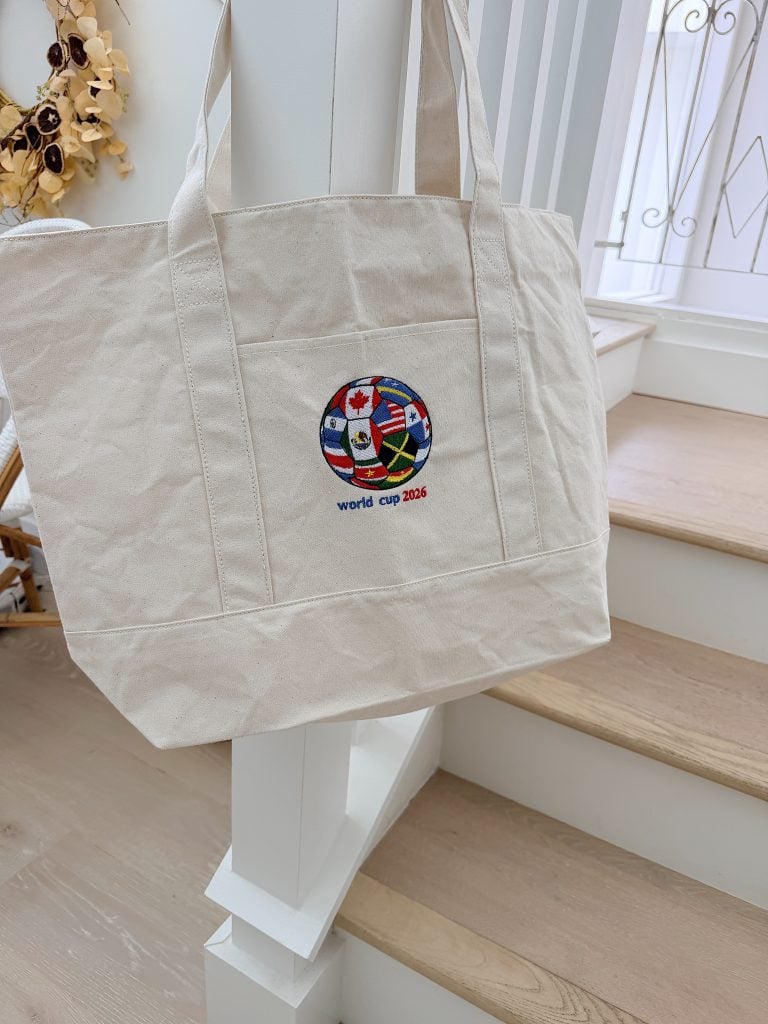 A beige canvas tote bag hangs on a white staircase railing, featuring a World Cup 2026 logo with various national flags.
