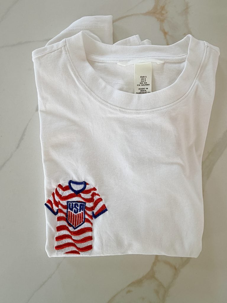 A folded white T-shirt with a small embroidered red and white striped jersey patch featuring a "USA" shield on the front.