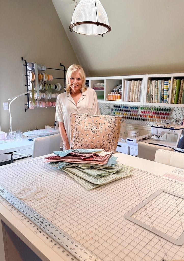 A woman stands in a craft room behind a table with folded fabrics and a patterned storage bin; shelves of fabric, ribbons, and spools are visible in the background.