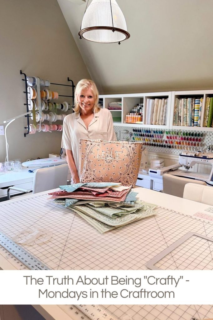 Woman stands in a well-organized craft room behind a table with stacked patterned fabrics; shelves with supplies are visible in the background.