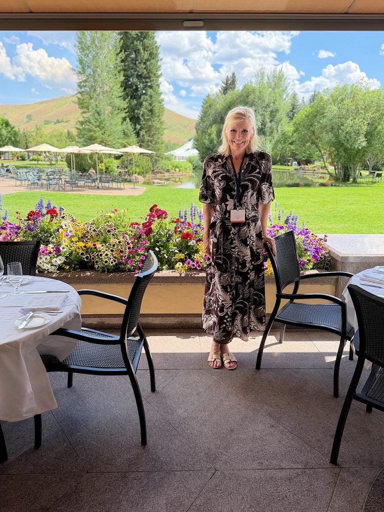 A woman in a patterned dress stands by a table on a covered patio, with colorful flowers and a green lawn in the background under a partly cloudy sky.