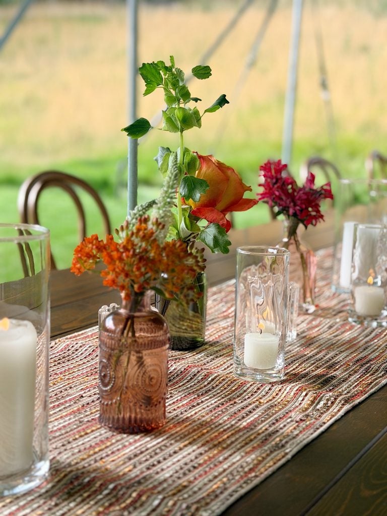 A wooden table with a woven runner, glass vases holding flowers and greenery, and glass candle holders with white candles.