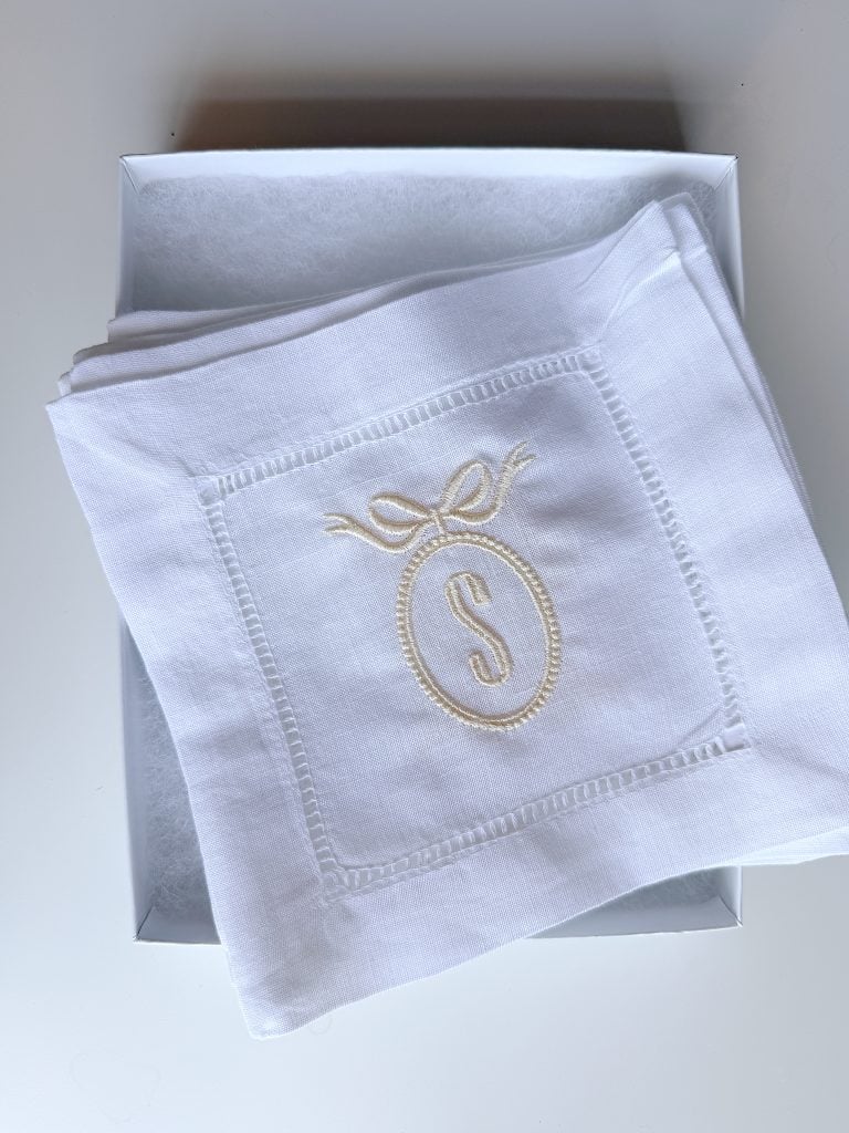 White linen napkin with an embroidered letter "S" inside an oval with a bow, neatly folded in a white box.